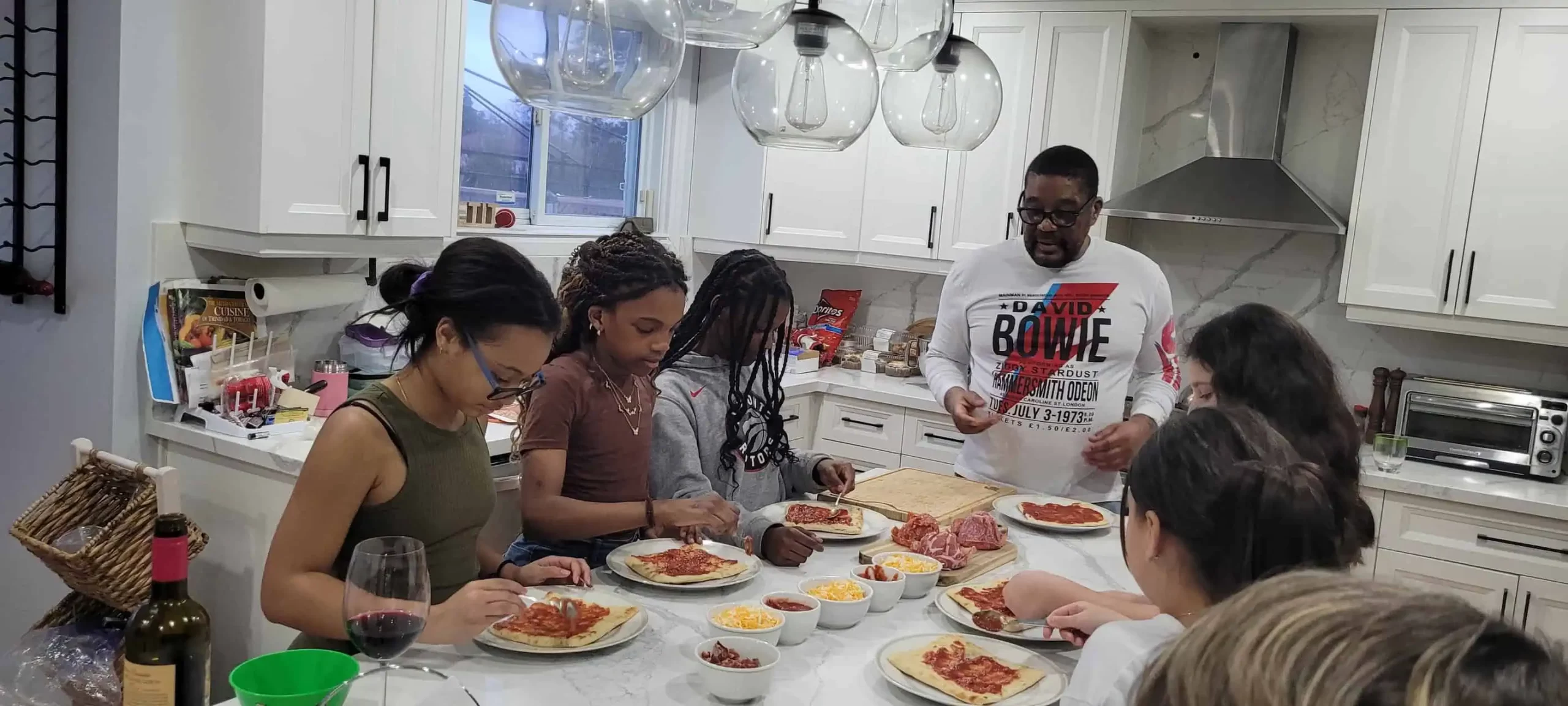 A group of people are gathered around a kitchen island making pizzas. There are various pizza toppings and ingredients on the counter. A man is instructing while others are placing toppings on their pizzas, showcasing how effective event management can turn any gathering into an engaging activity.