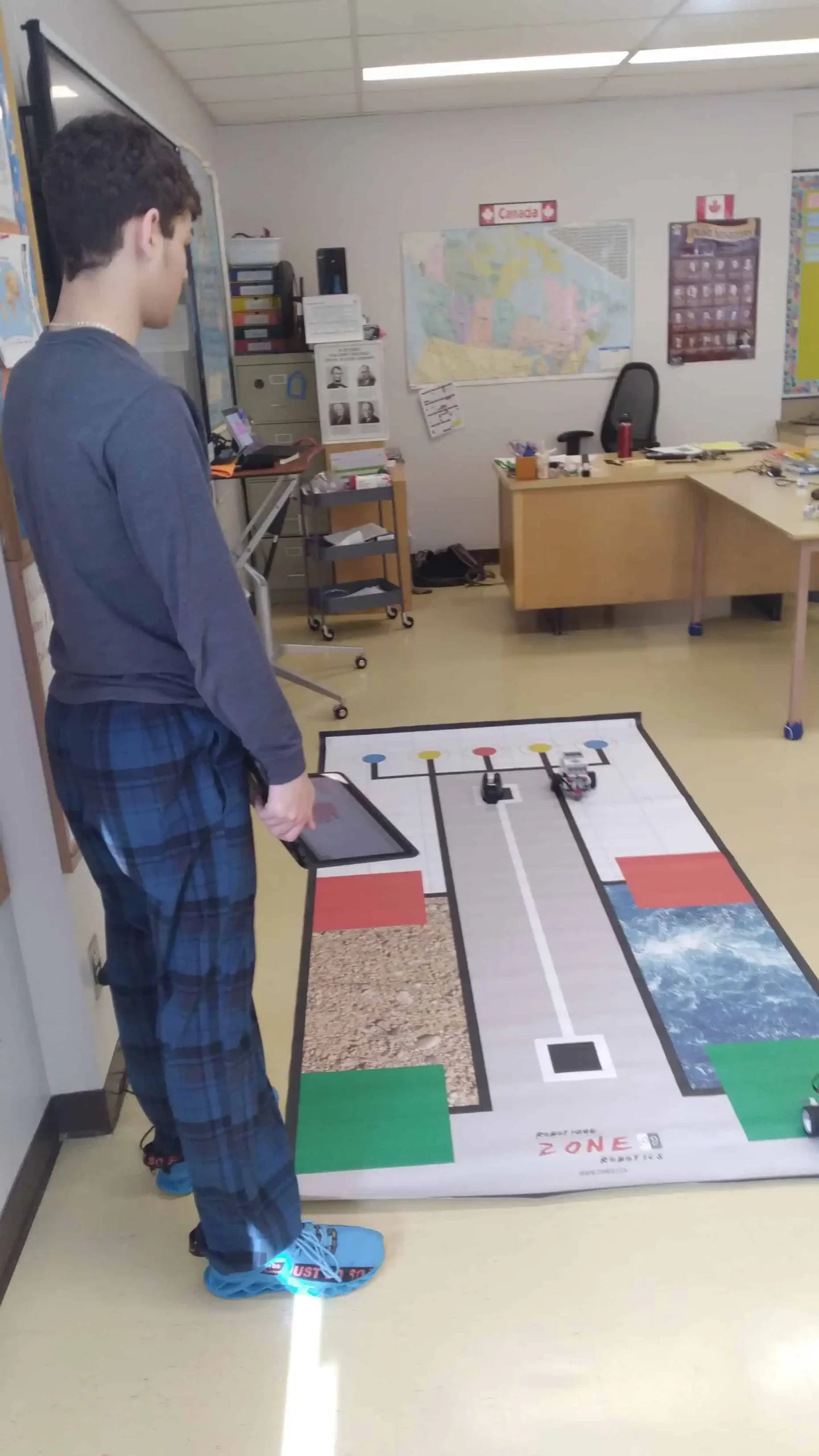 A person stands in a classroom holding a tablet, observing a mat on the floor designed with various paths and colored zones, possibly for a robotic programming activity. This scene could easily be part of an A Blue Door event marketing initiative focused on educational technology.