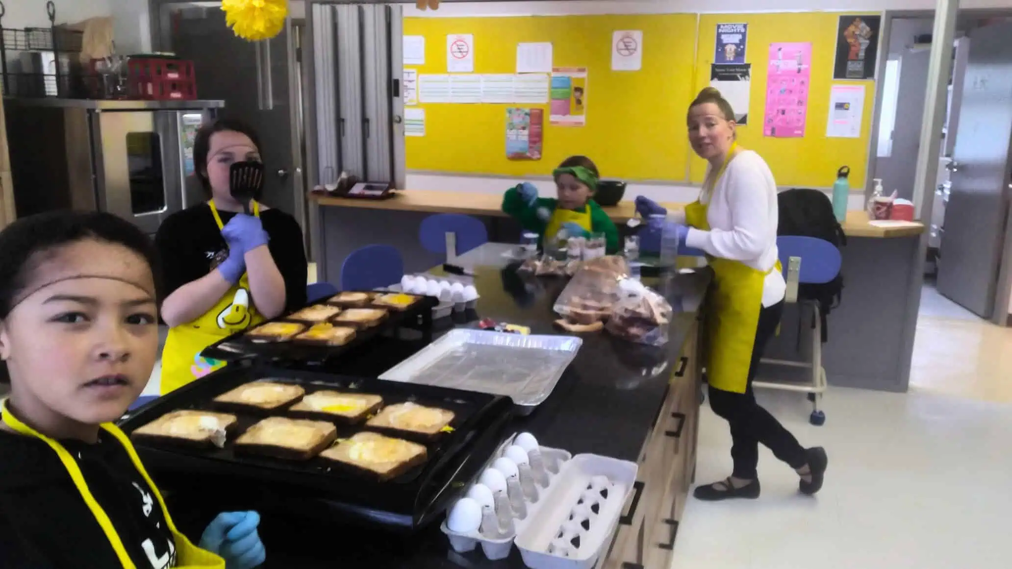 Children and an adult prepare meals in a kitchen, wearing aprons and gloves. Food trays, bread, eggs, and utensils are on the counter. A yellow bulletin board with posters is visible in the background. Through A Blue Door beside them, more students arrive to join the fun.