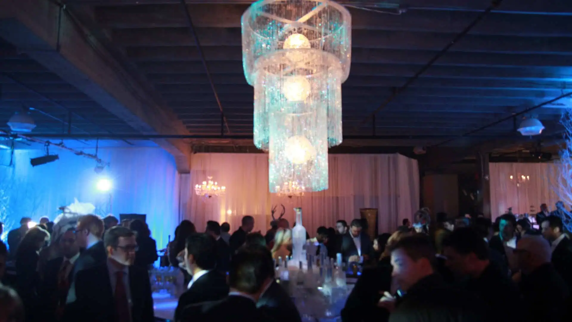 A large chandelier with blue lighting hangs above a crowd of people dressed in formal attire at an indoor event flawlessly organized by A Blue Door, showcasing their impeccable event planning skills.