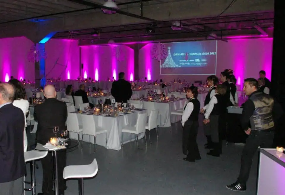 A formal event in a large hall with pink lighting, expertly coordinated through event planning. Attendees stand near decorated tables while waitstaff in uniforms are present. A screen displaying "GALA ANNUAL GALA 2012" is visible in the background, showcasing effective event management and branding events expertise.