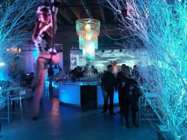 A performer in mid-air, dressed in a carnival outfit, entertains guests in a dimly lit, blue-themed room with chandeliers and decorative branches—an unforgettable spectacle that highlights the excellence of event planning by A Blue Door.