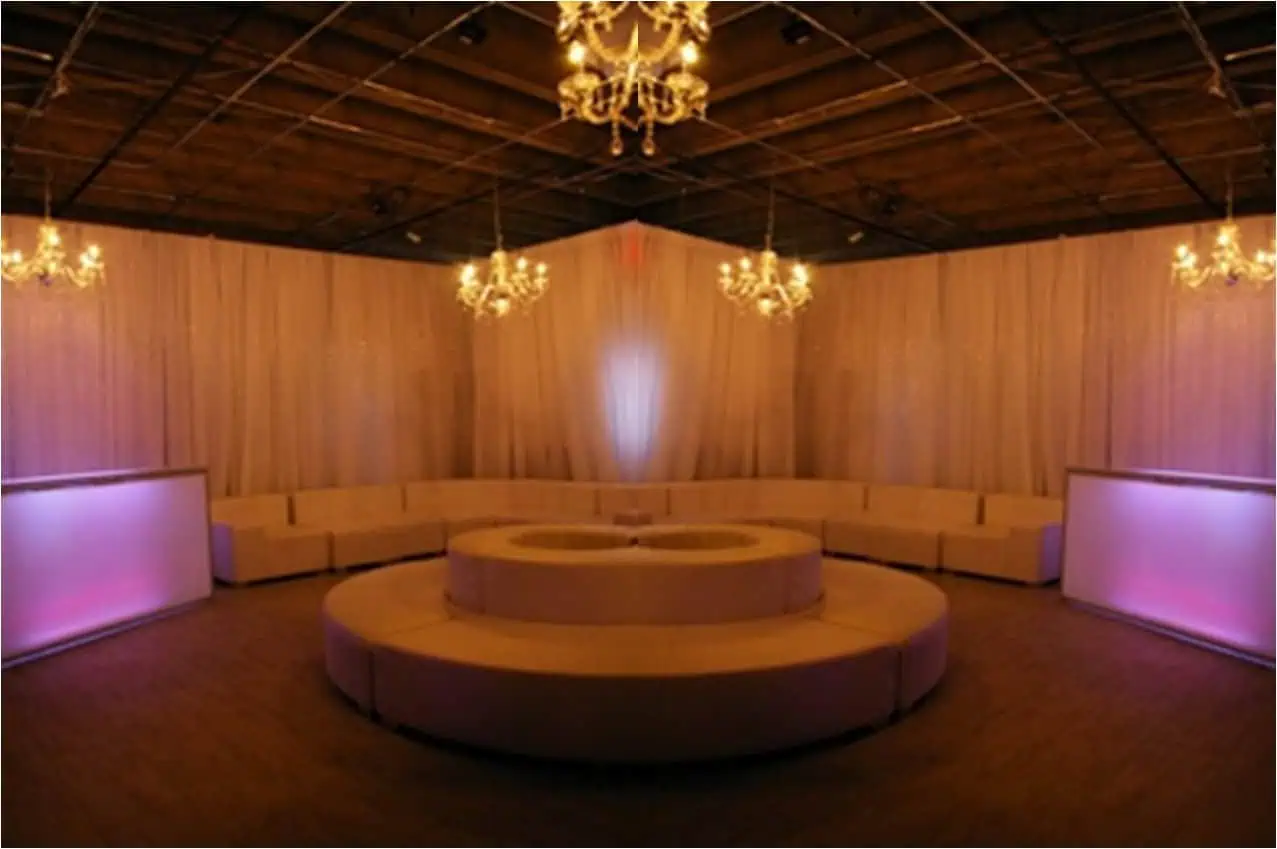 Dimly lit room with chandeliers, draped curtains, and a circular seating arrangement around a central fixture. LED lights provide a soft pink and purple hue, perfect for an event planned by A Blue Door.