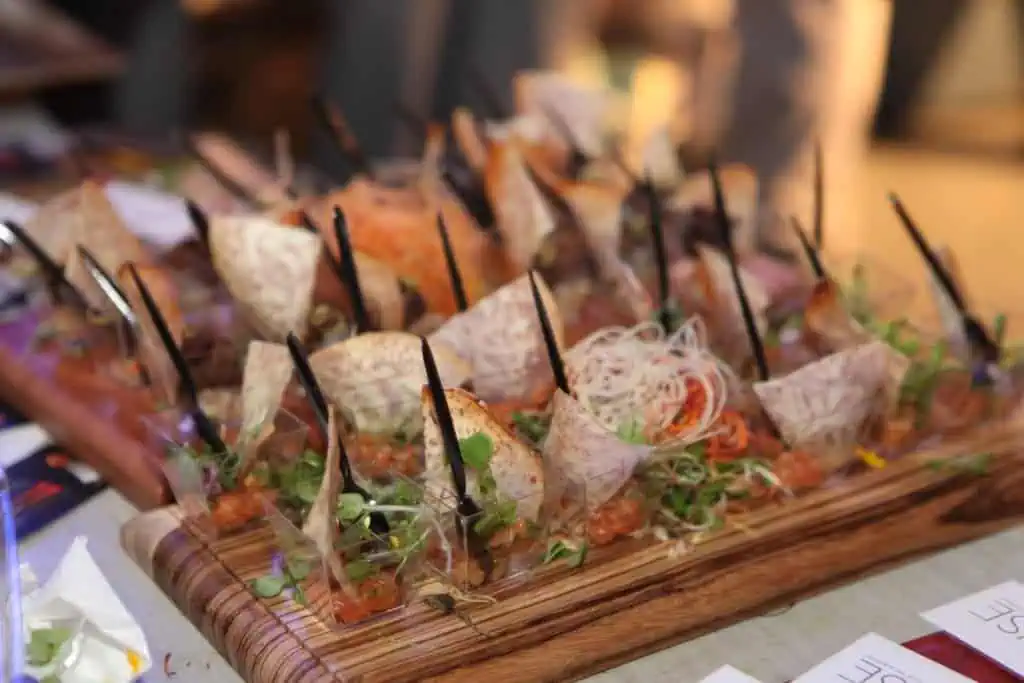 A wooden platter with assorted appetizers and garnishes, featuring ingredients like tortilla chips, vegetables, and sprouts, is elegantly displayed for an event—ideal for showcasing your event planning expertise.